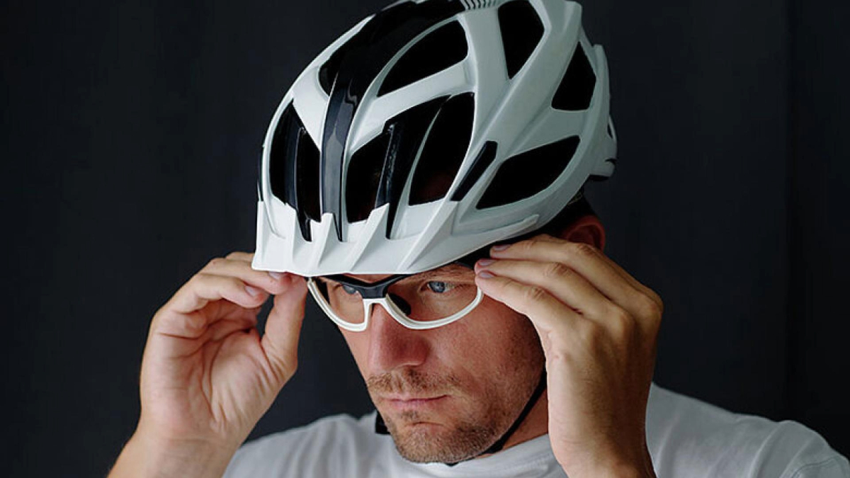 Person adjusting cycling glasses while wearing a white bike helmet against a dark background.