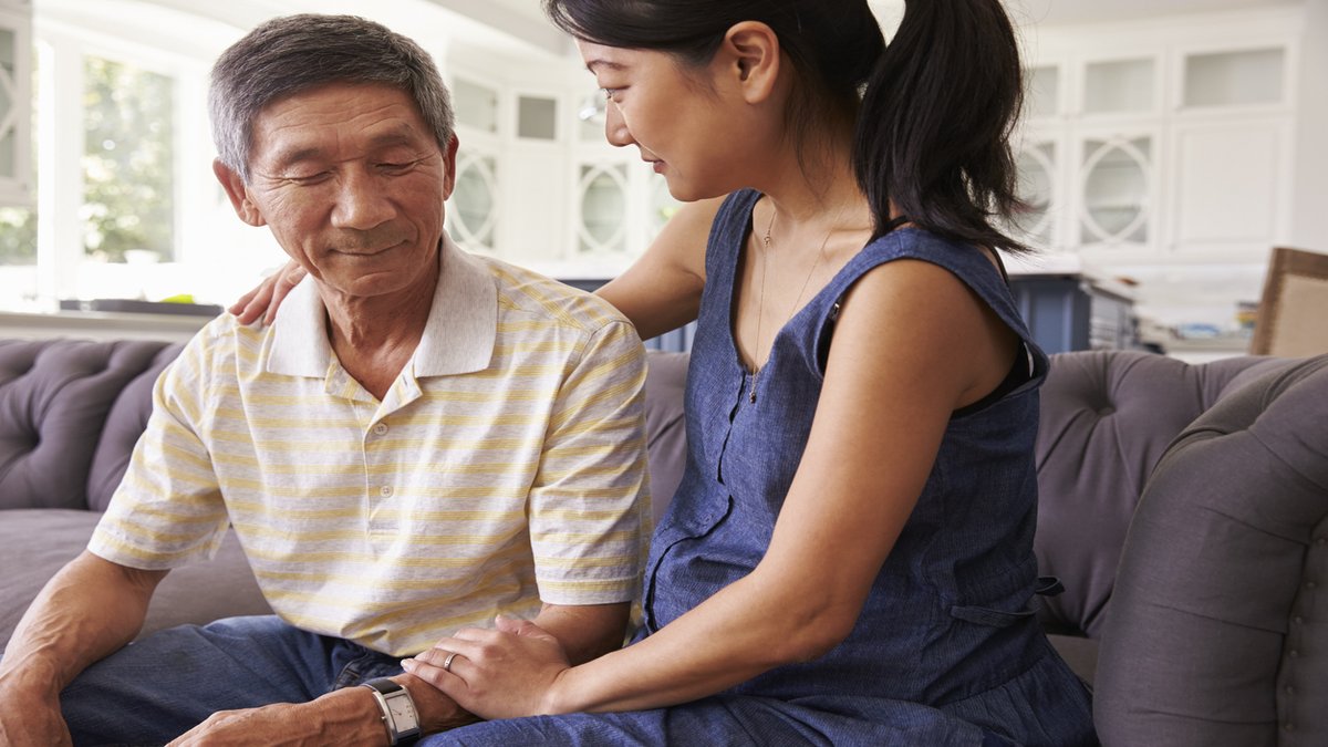 A young woman sits beside an older man on a couch, gently placing her hand on his shoulder in a comforting gesture. The scene reflects caregiving, support, and planning for dementia care in a warm, home environment.