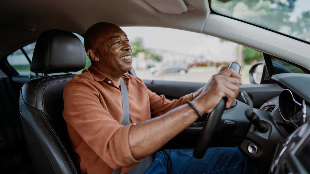 A smiling middle-aged man wearing a brown shirt is driving a car. He is looking ahead with a cheerful expression as he holds the steering wheel, enjoying the drive.