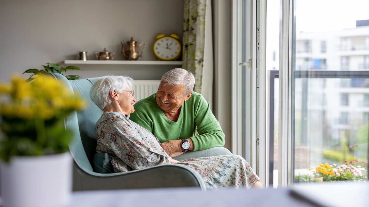 An elderly couple sitting together in a cozy living room near a large window. The man, dressed in a green sweater, is smiling warmly at the woman, who is seated comfortably in a chair. They appear to be sharing a moment of affection and conversation in a peaceful, homey environment.