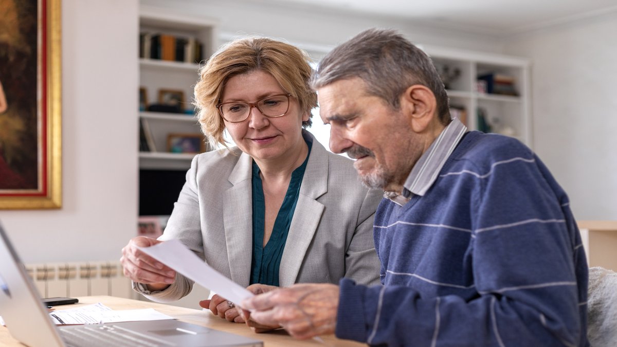 A middle-aged woman in glasses and a gray blazer is sitting next to an elderly man in a blue sweater. They are reviewing documents together at a table, with a laptop open in front of them, in a bright, well-lit living room.