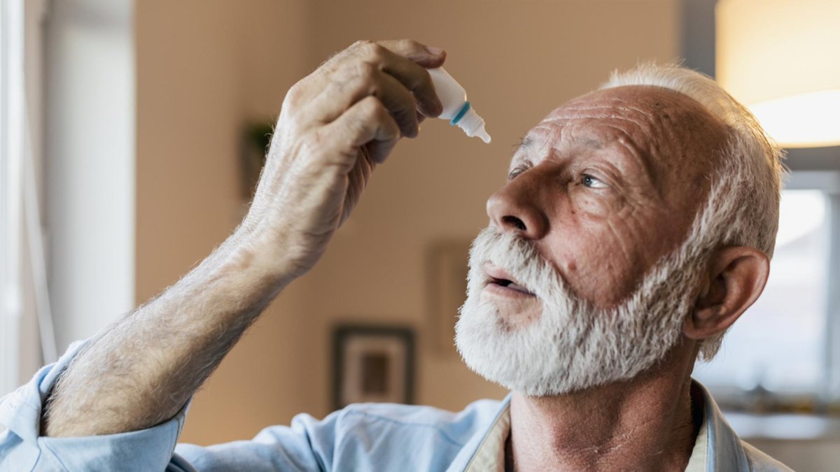 An elderly man with a white beard is tilting his head back and applying eye drops at home.