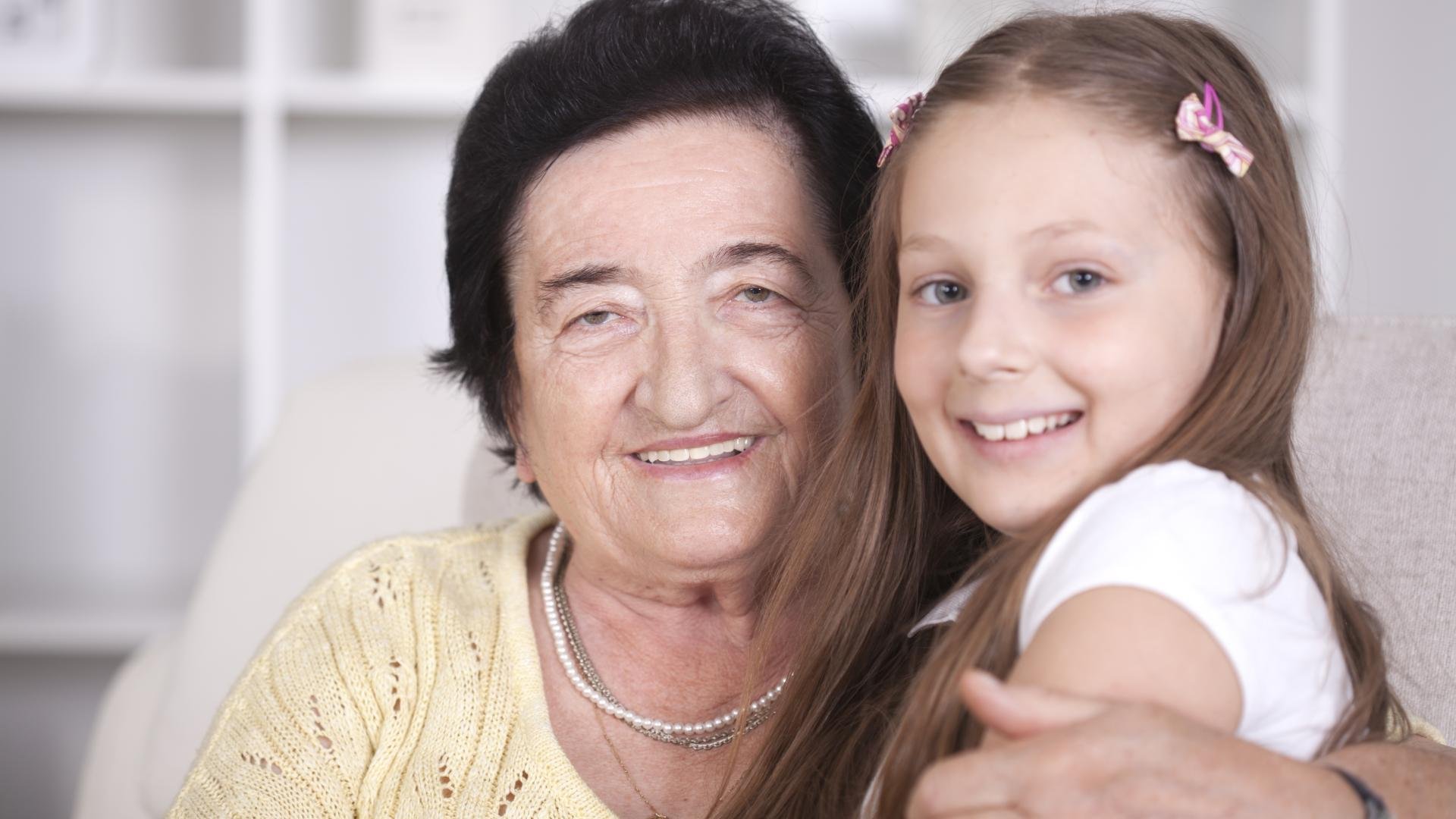 An elderly woman with short dark hair and a warm smile sits closely with a young girl who has long brown hair, wearing a white shirt and hair clips. They are both smiling and embracing, creating a sense of warmth and connection in a bright indoor setting.