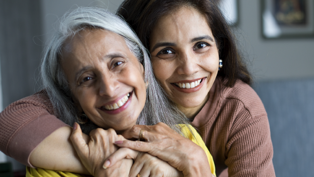 Two smiling individuals, one older and one younger, embracing affectionately in a cozy setting.