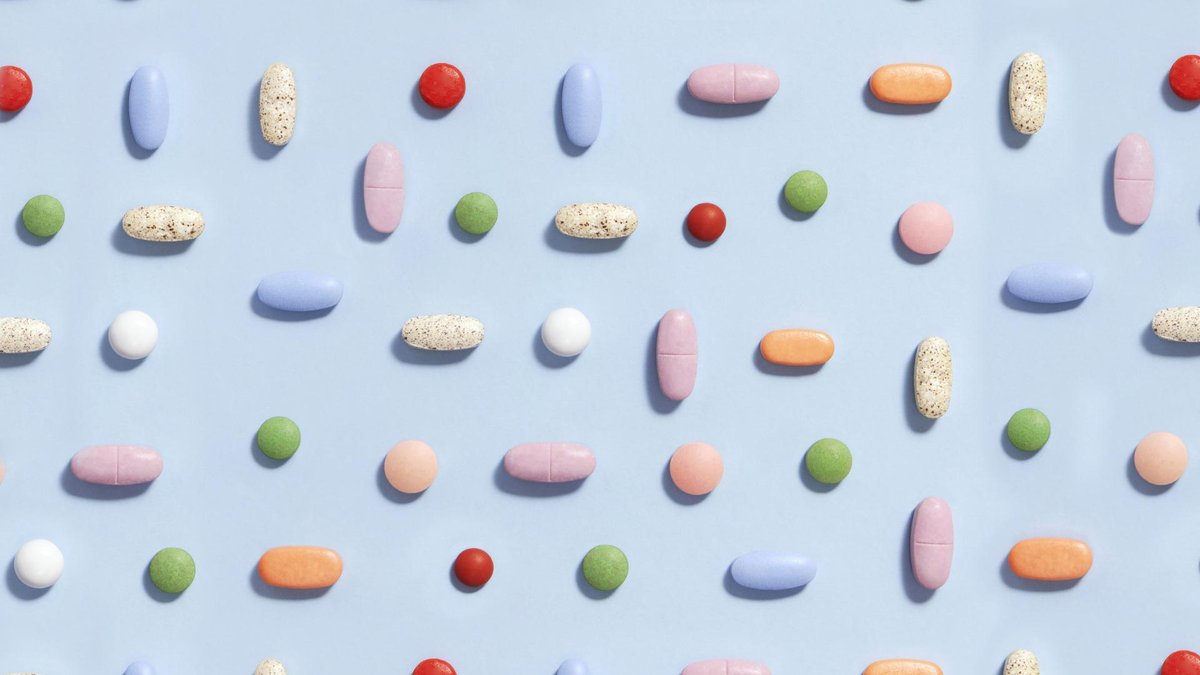 A flat lay image of various colorful pills and tablets arranged neatly on a light blue background. The pills vary in shape, size, and color, including red, green, orange, pink, blue, white, and speckled designs.