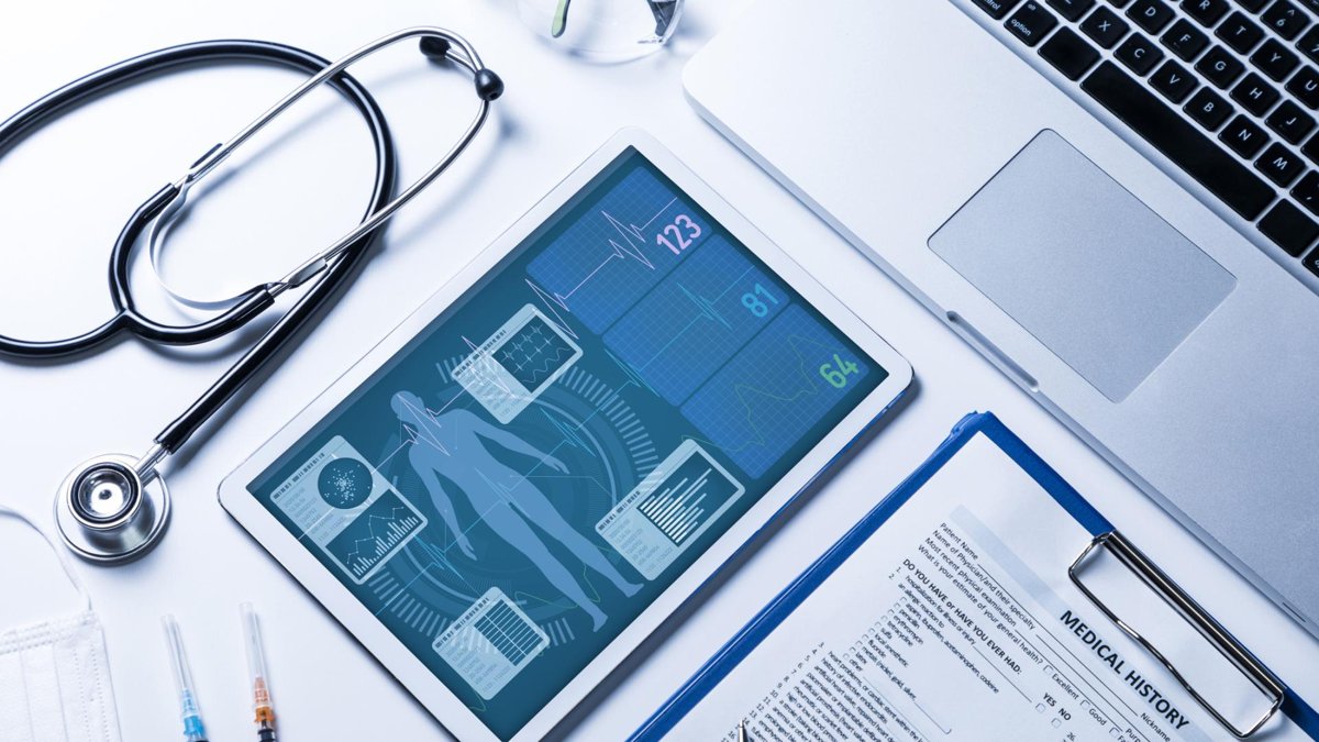 A medical setup with a stethoscope, a tablet displaying digital health data, a laptop, and a medical history form on a clipboard, representing modern healthcare technology and patient records