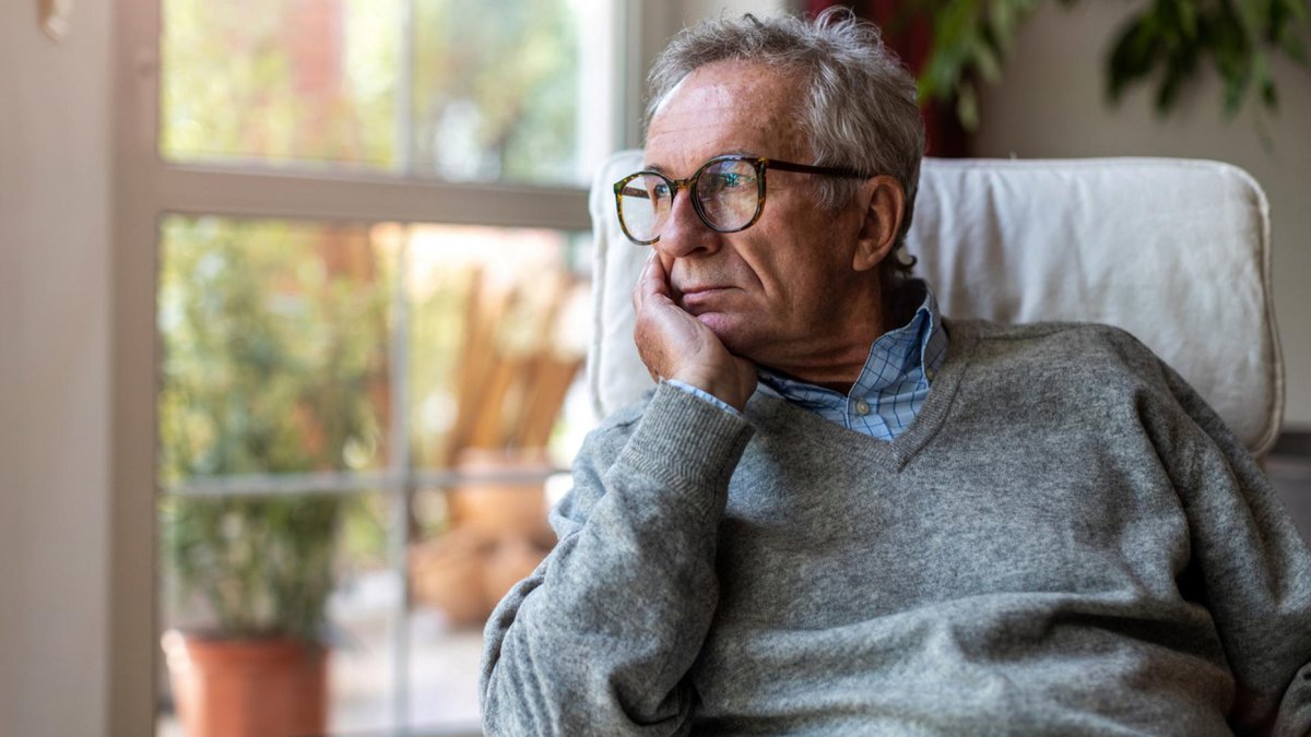 Senior man with glasses looks out window thoughtfully.