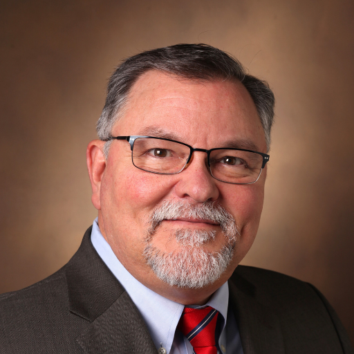 Man with glasses and a gray beard, wearing a suit and red tie, smiling at the camera.