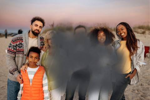 A group photo of a family standing on a beach at sunset. The family consists of a man in a sweater, a young boy in an orange vest, an older woman, a younger woman with curly hair, and a woman with long braids. The faces of the older woman and the younger woman with curly hair are blurred.