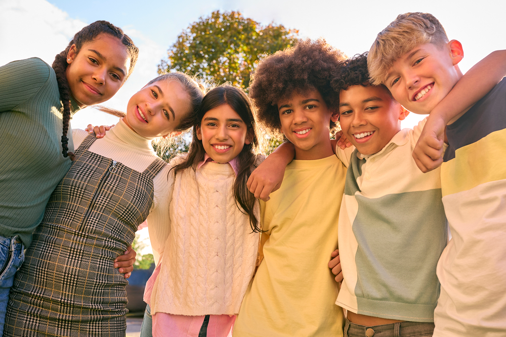 A group of six children of diverse backgrounds smiling and standing close together with their arms around each other. They are outdoors on a sunny day with a tree and blue sky in the background.