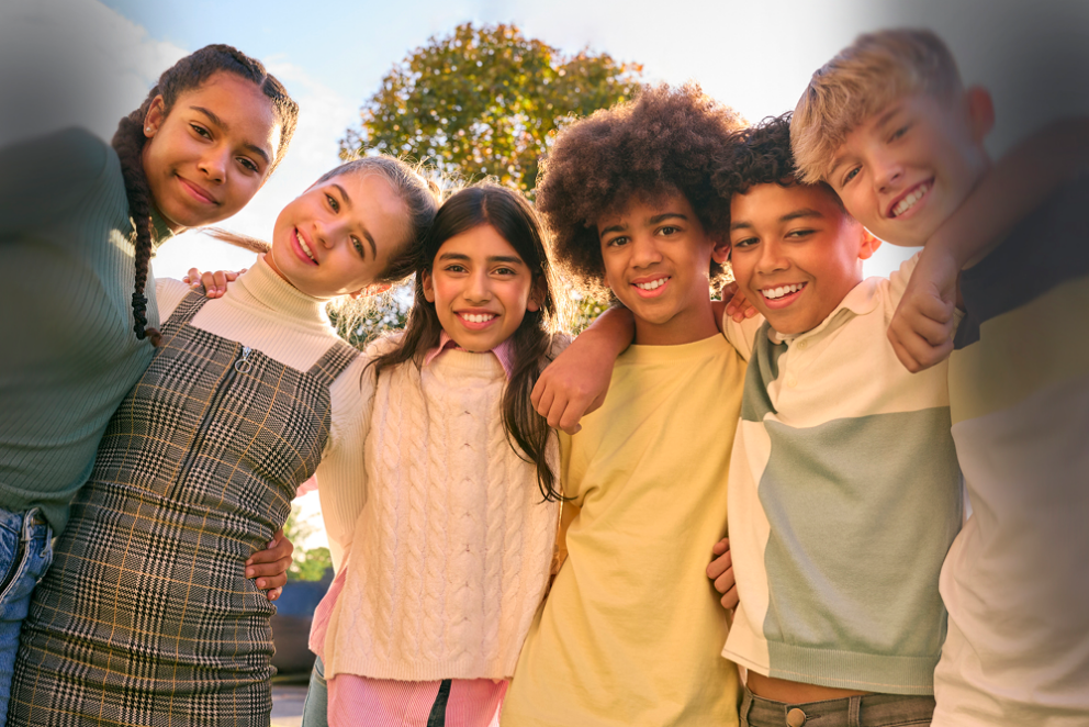 A group of six children of diverse backgrounds smiling and standing close together with their arms around each other. They are outdoors on a sunny day with a tree and blue sky in the background, with a vignette effect around the edges.