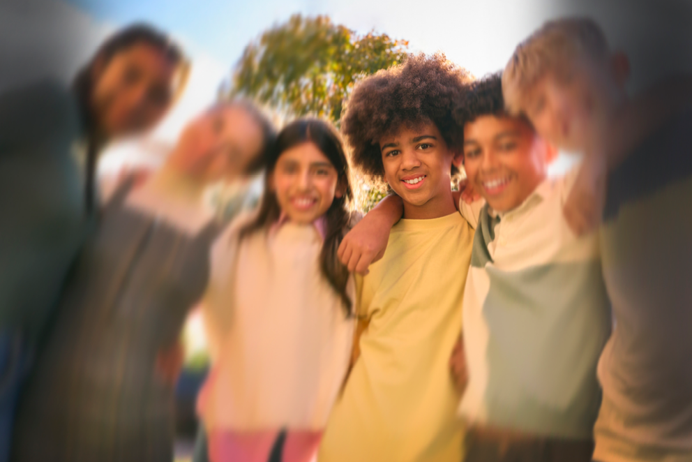 A group of six children of diverse backgrounds smiling and standing close together with their arms around each other. They are outdoors on a sunny day with a tree and blue sky in the background, with a strong blur effect around the edges, focusing on the center child.