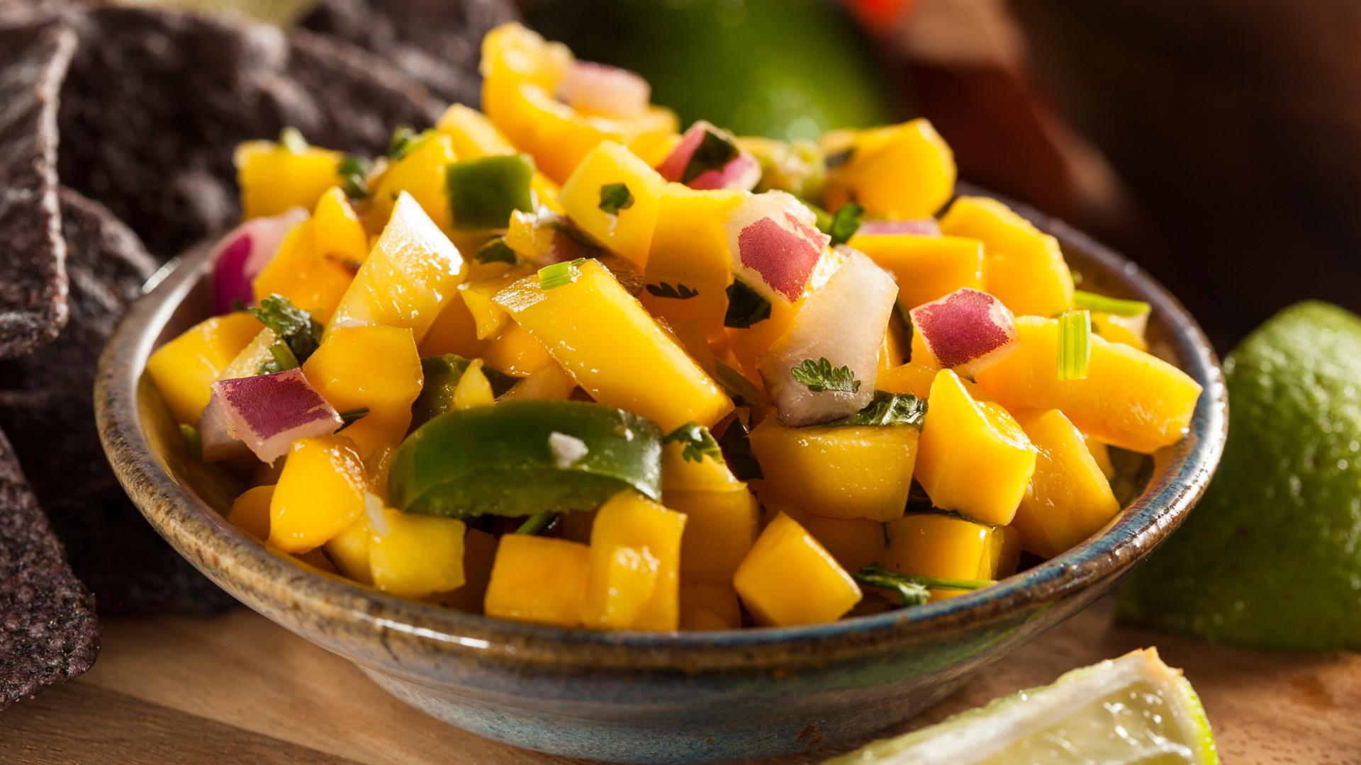 A bowl of mango salsa with diced mango, green bell peppers, red onions, and cilantro, accompanied by lime wedges and tortilla chips in the background.