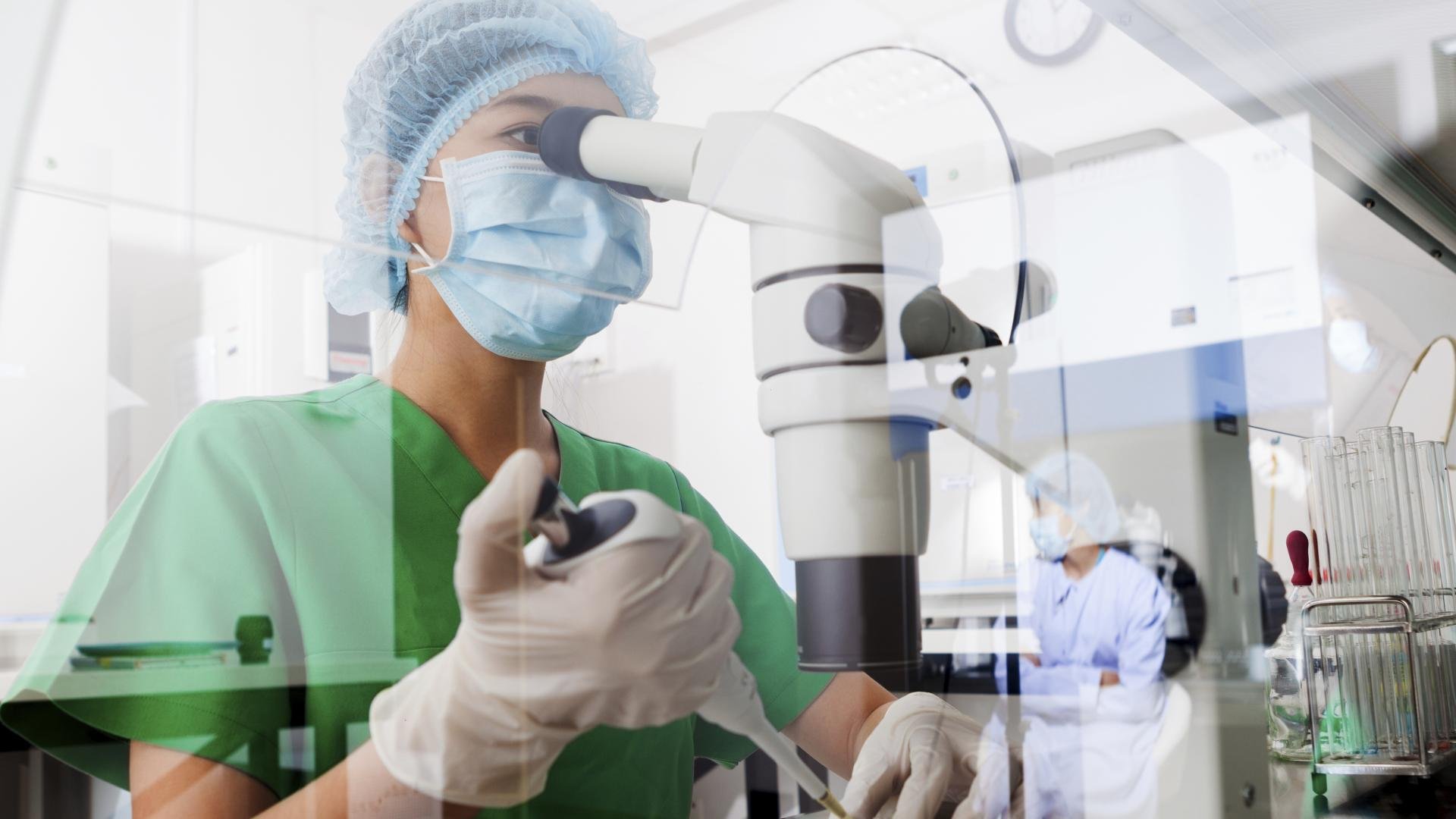 A researcher in a lab wearing a mask, gloves, and a green scrub suit, using a microscope while working with lab equipment and test tubes.