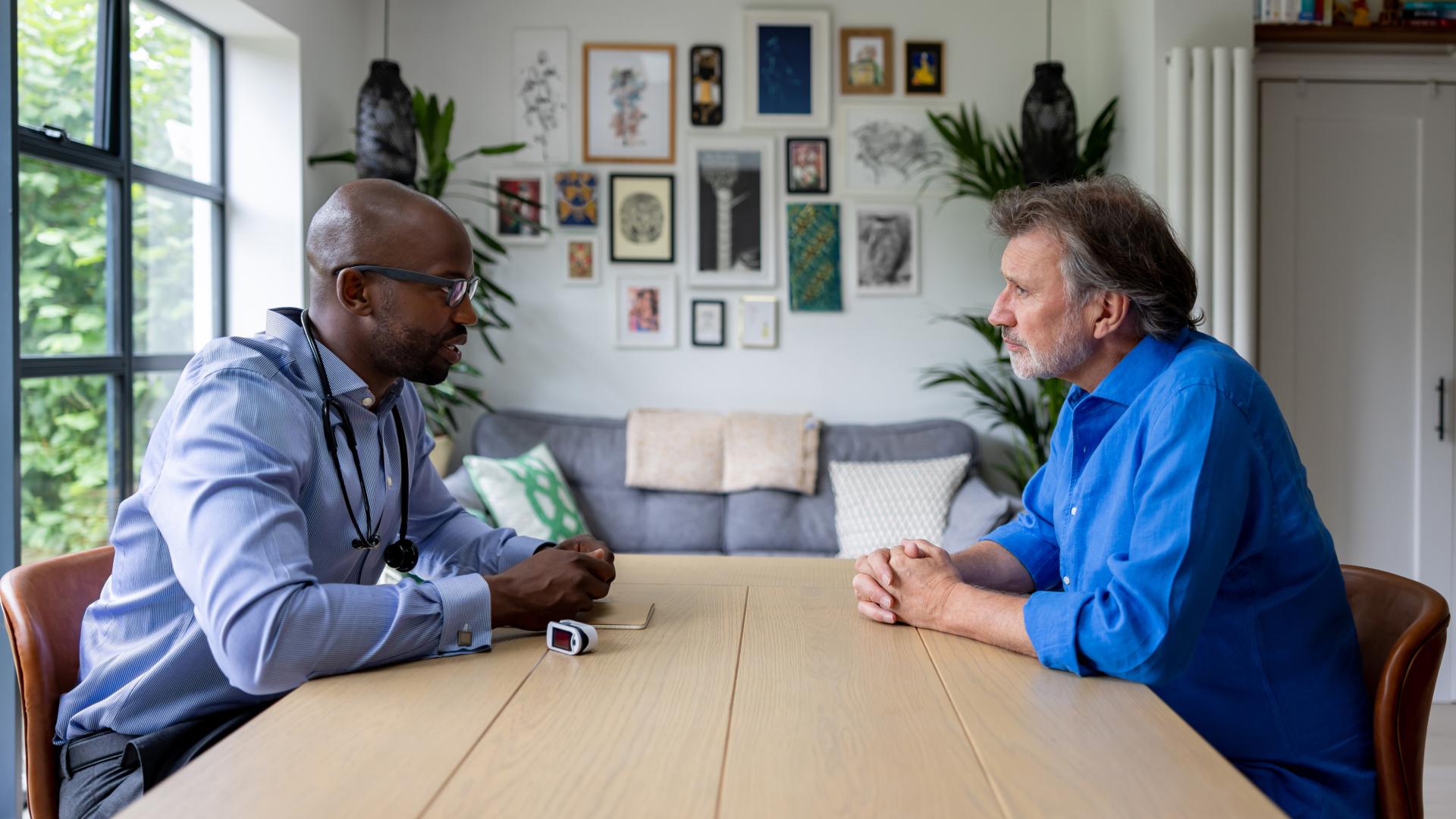 Two men dressed in business casual clothes sit across from each other at a table. The one on the left has a stethoscope around his neck.