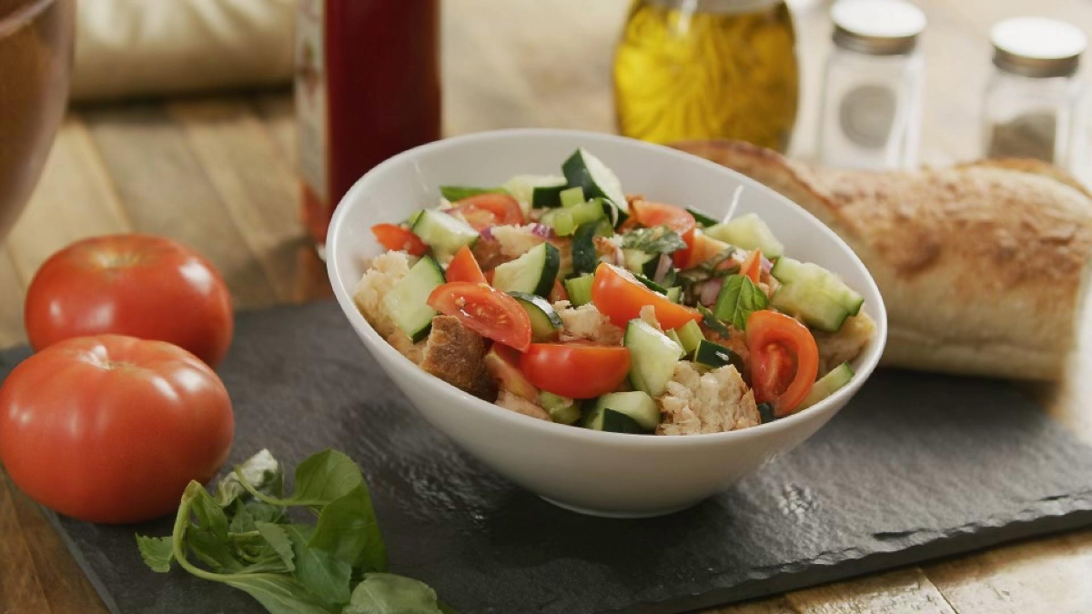 A bowl of fresh Tuscan bread salad, also known as Panzanella salad, containing tomatoes, cucumbers, and croutons, served with a side of bread and garnished with herbs, placed on a wooden table with tomatoes, salad leaves, and condiments around it.