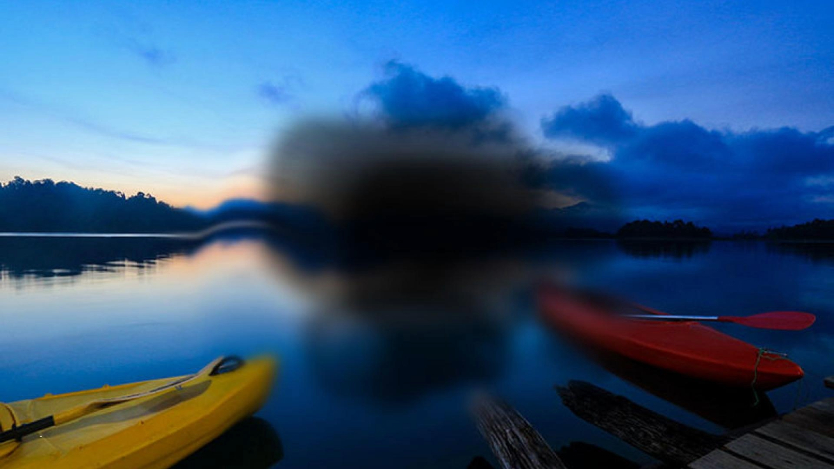 Three kayaks, one red and two yellow, are lined up on a wooden dock beside a calm blue lake during twilight, with a blurred area in the center of the image.