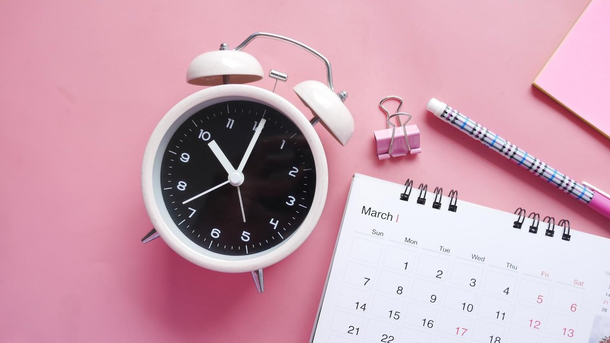 A top view of a desktop with a retro-style alarm clock, a March calendar, a pink paper clip, and a patterned pen placed on a pink background.