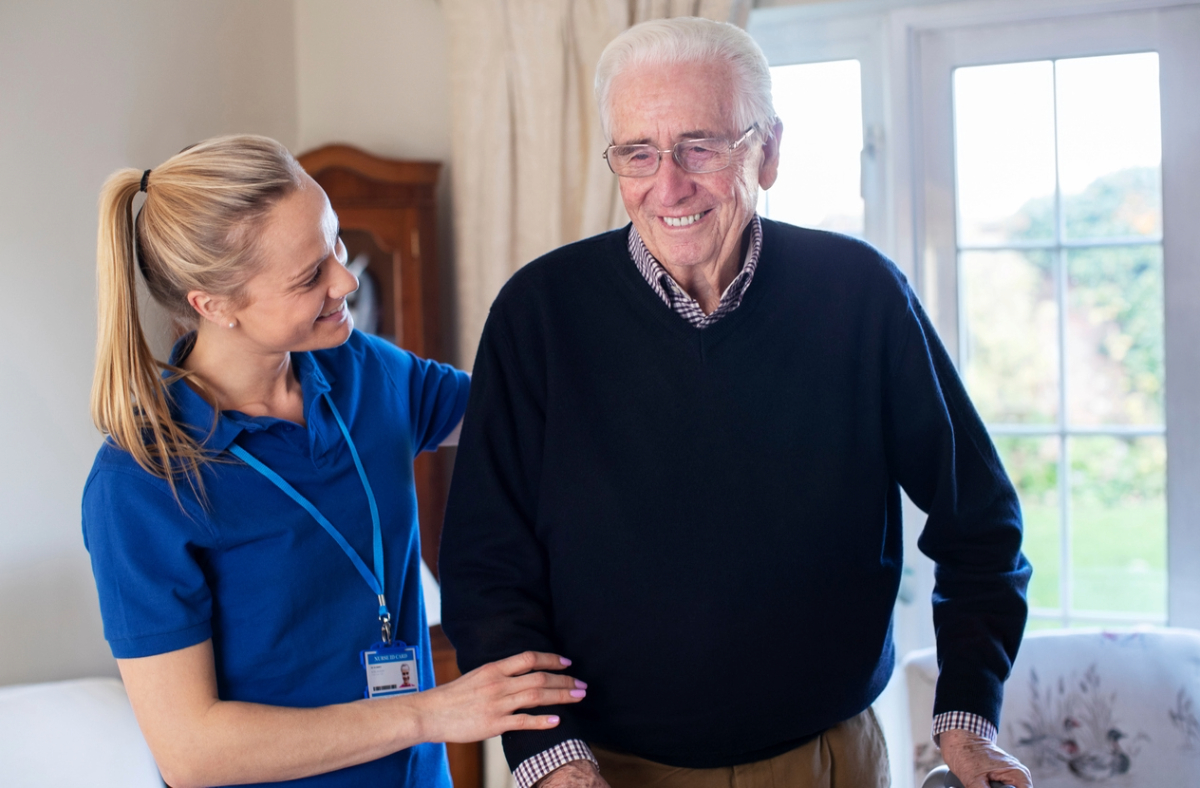 A healthcare worker in a blue uniform is assisting an elderly person wearing glasses and a sweater, standing in a well-lit room.