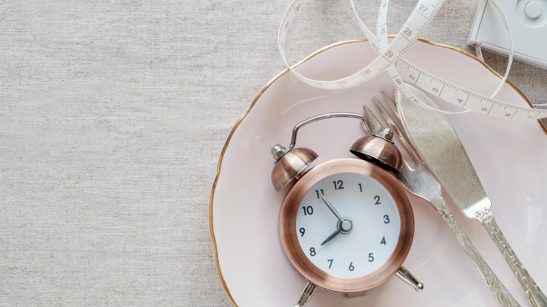 a clock, a tape measure, and utensils sit on a pink plate with a linen background