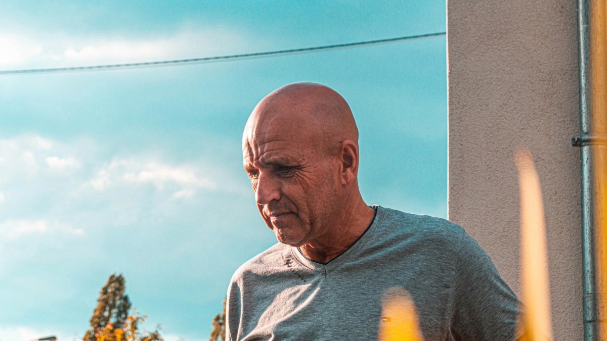 A person with a bald head wearing a grey shirt, looking downward with a thoughtful expression, outdoors under a blue sky.