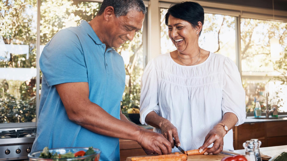 Two people are joyfully preparing food together in a sunlit kitchen, chopping vegetables on a wooden countertop.