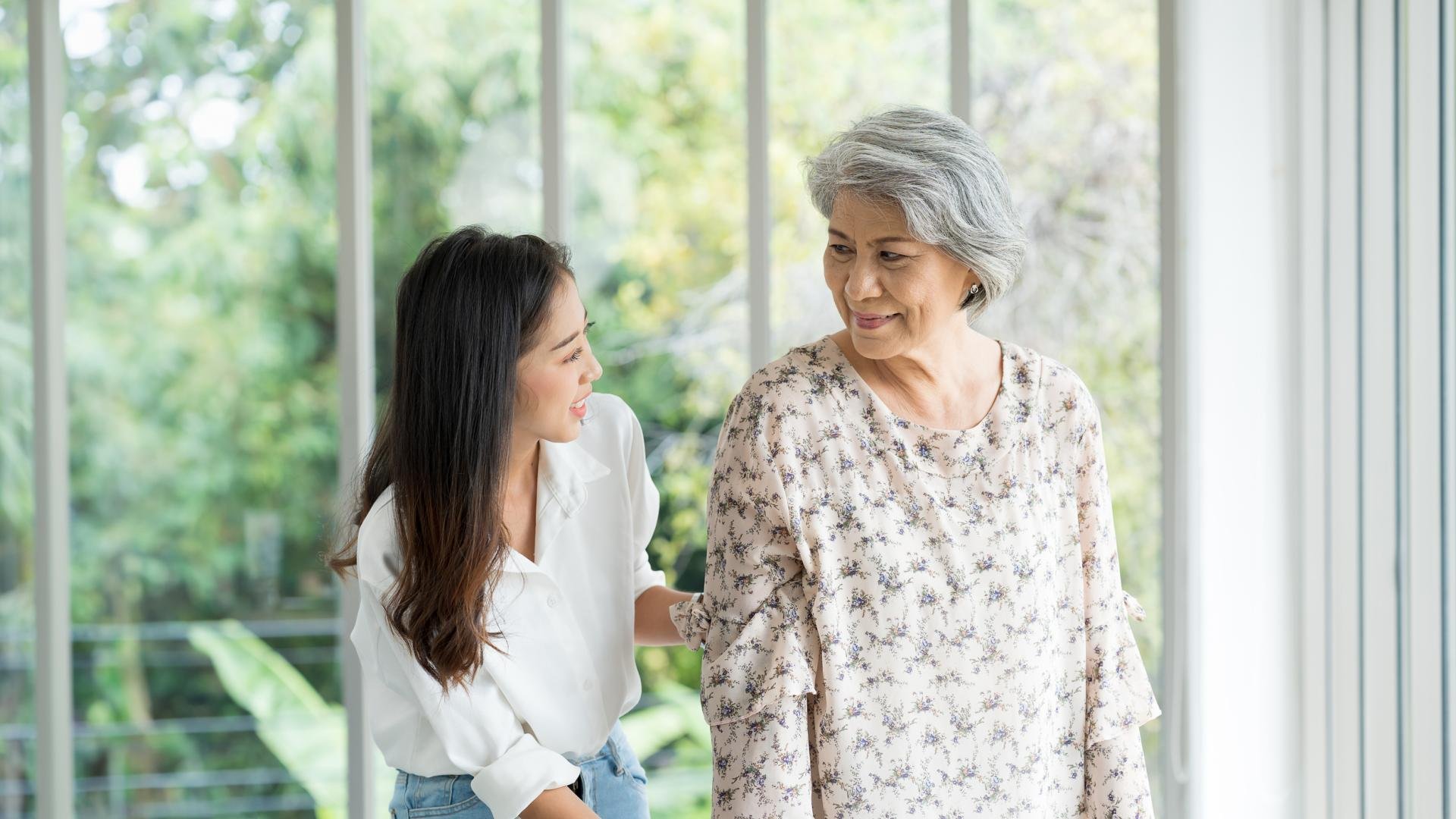 A young woman assists her elderly mother, both smiling warmly at each other while standing in a bright room with large windows and greenery in the background.