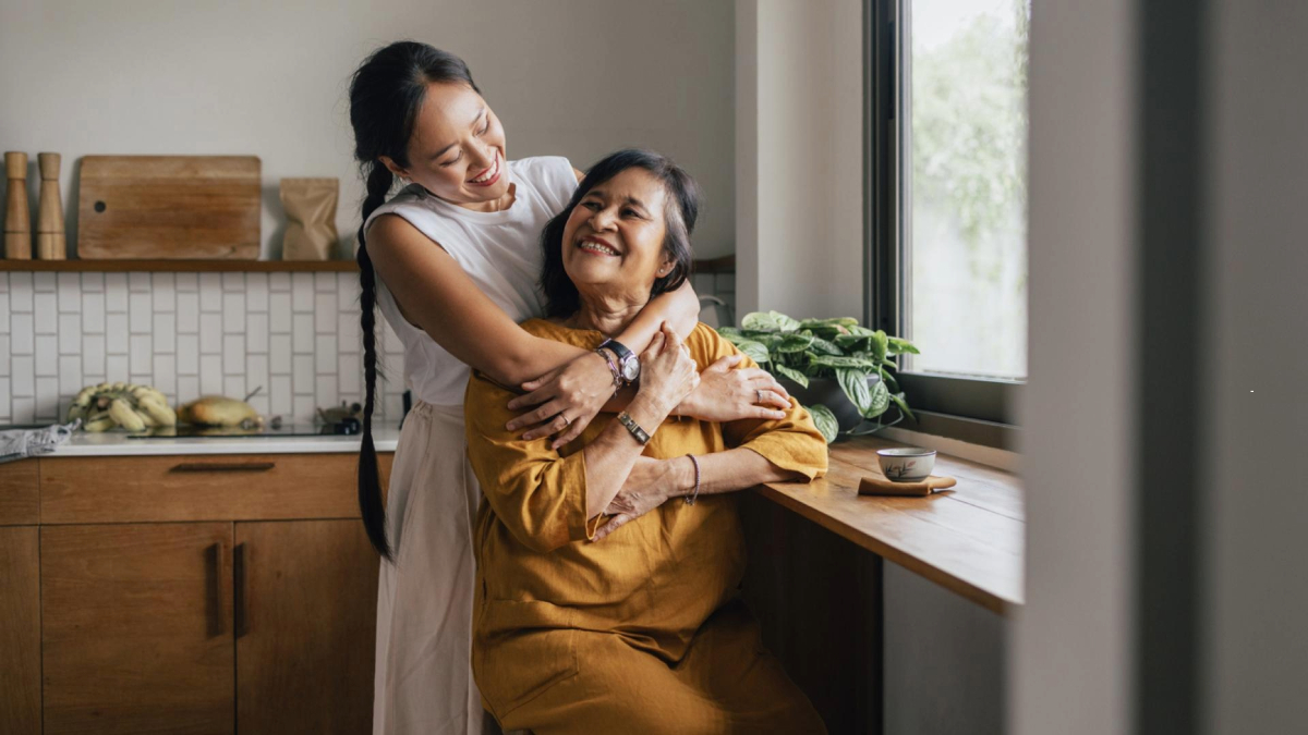 Two adults are sharing a hug in a kitchen setting, with one person standing and embracing the other who is seated. The kitchen has a modern decor with visible vegetables and a window.