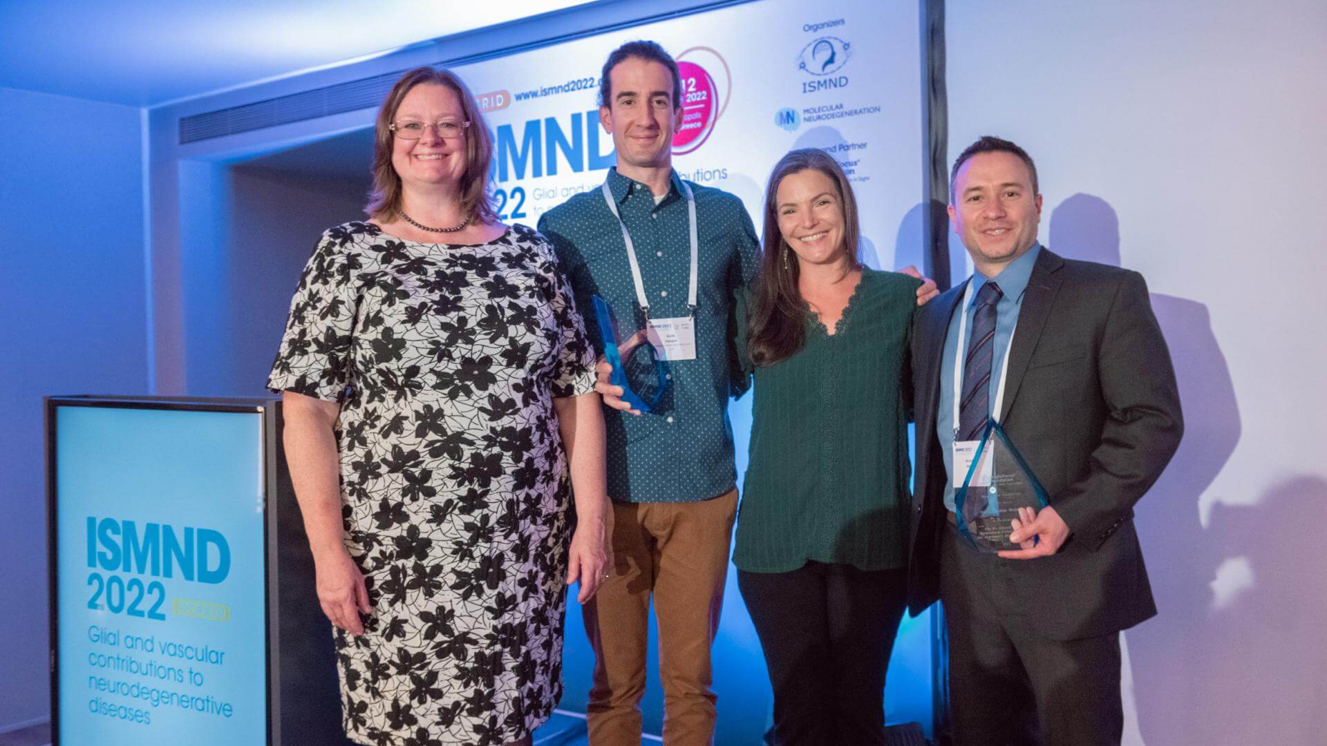 Diane Bovenkamp, Keith Hengen Sharyn Rossi, and Brandon Holmes group picture in front of podium with the ISMIND 2022 logo on it in blue text.