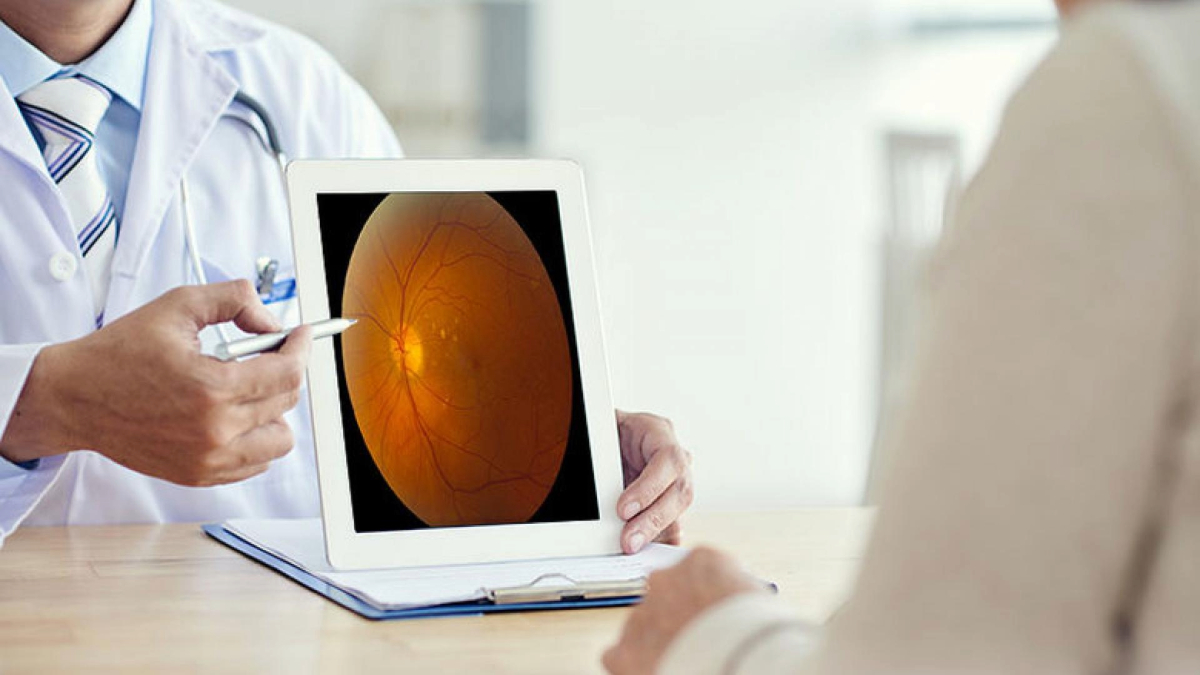 A healthcare professional is showing a patient an eye exam result on a tablet screen displaying an image of a retina. They are seated at a desk in a clinical setting.