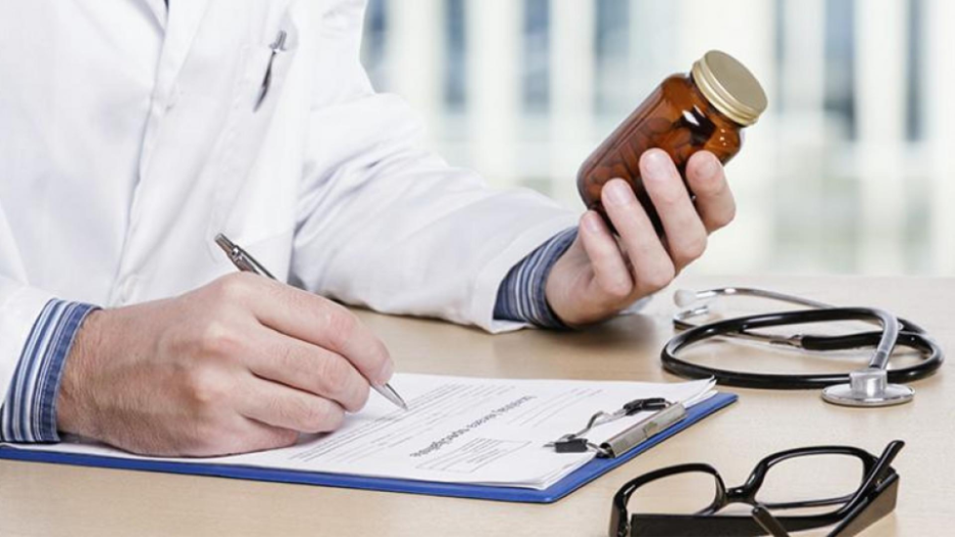 A photograph of someone wearing a white doctor's coat over a blue and white striped long-sleeved shirt holding a dark glass bottle in one hand and a pen - writing on a piece of paper on a clipboard - in the other. Only part of their forearms and chest are visible. A stethoscope and glasses rest on the table near the clipboard.