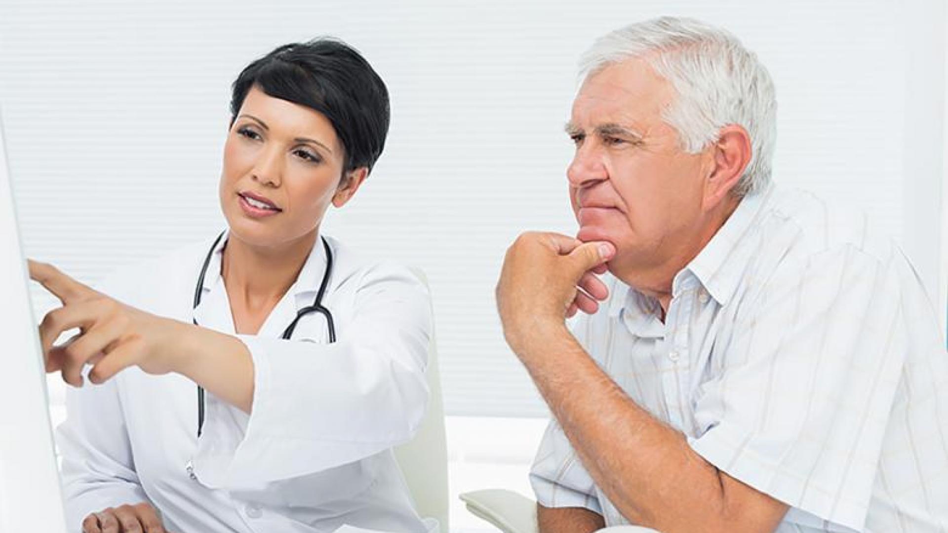 A woman wearing a white doctor's coat points at something outside of the image. The older man sitting next to her rests his hand on his chin and appears to be listening intently.