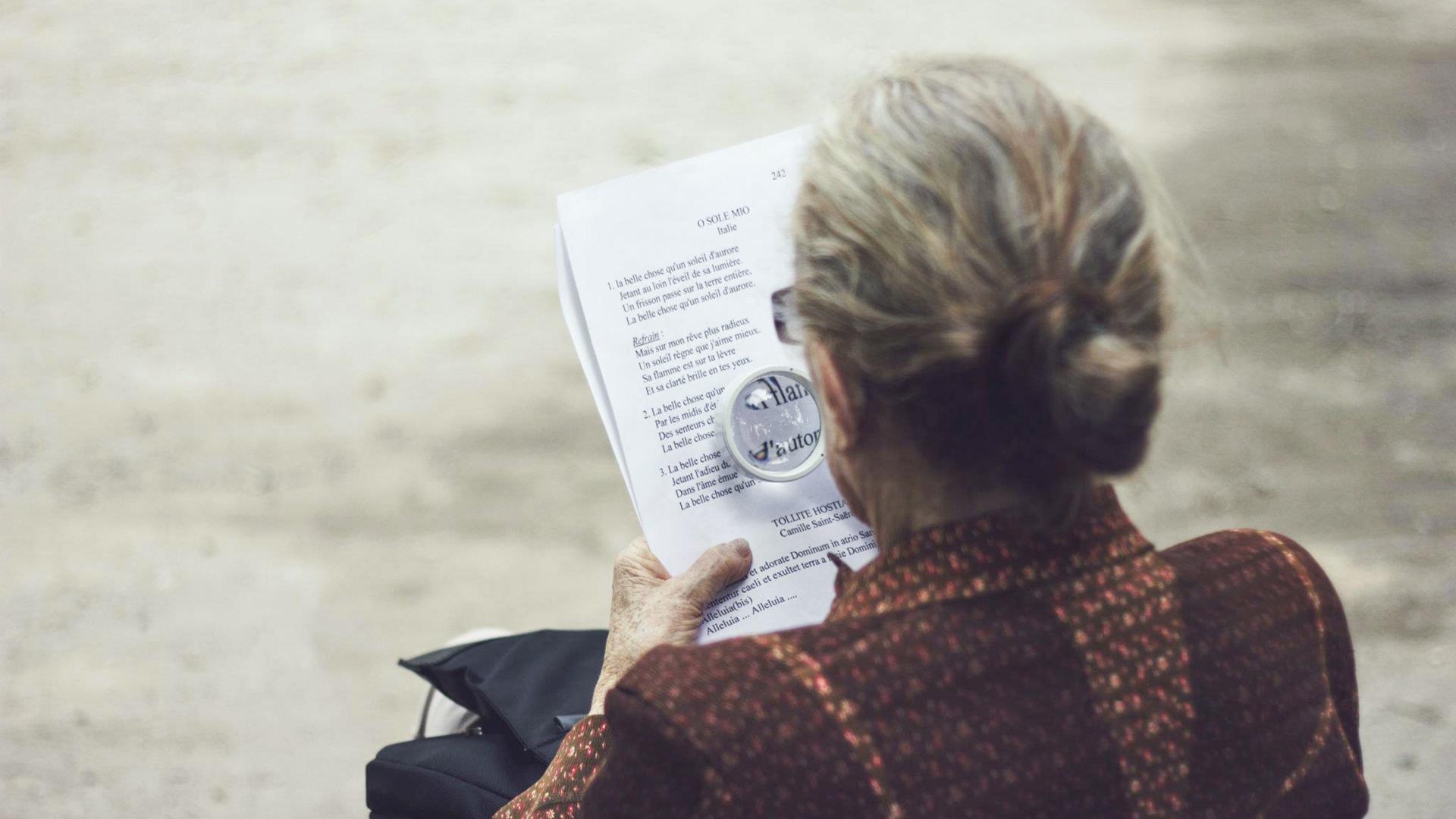 a photograph of an elderly woman from behind. She is using a magnifying glass to read something off a piece of paper.