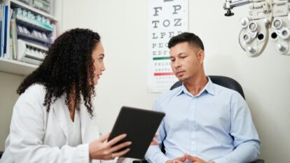 An eye doctor sitting in an exam room, holding a tablet and discussing something with a male patient. The patient is seated, looking attentively at the doctor, with an eye chart and medical equipment visible in the background.