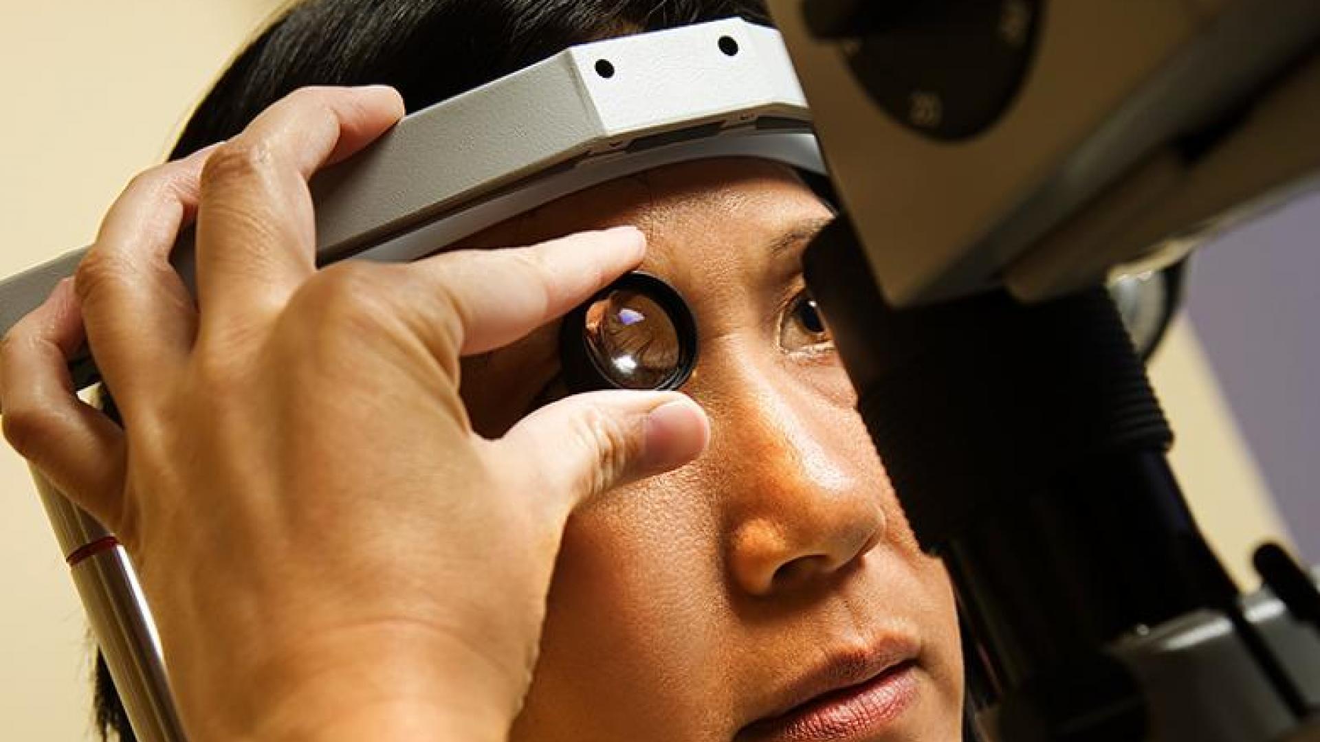 A close-up of a person undergoing an eye examination using a gonioscopy lens. The patient’s head is positioned on a diagnostic device while the examiner holds the lens near the eye to inspect the drainage angle for glaucoma or other eye conditions.