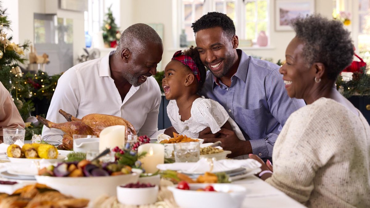 A joyful multigenerational family shares a holiday meal together. The group is smiling and laughing, gathered around a beautifully set dinner table with holiday decorations and a roasted turkey.