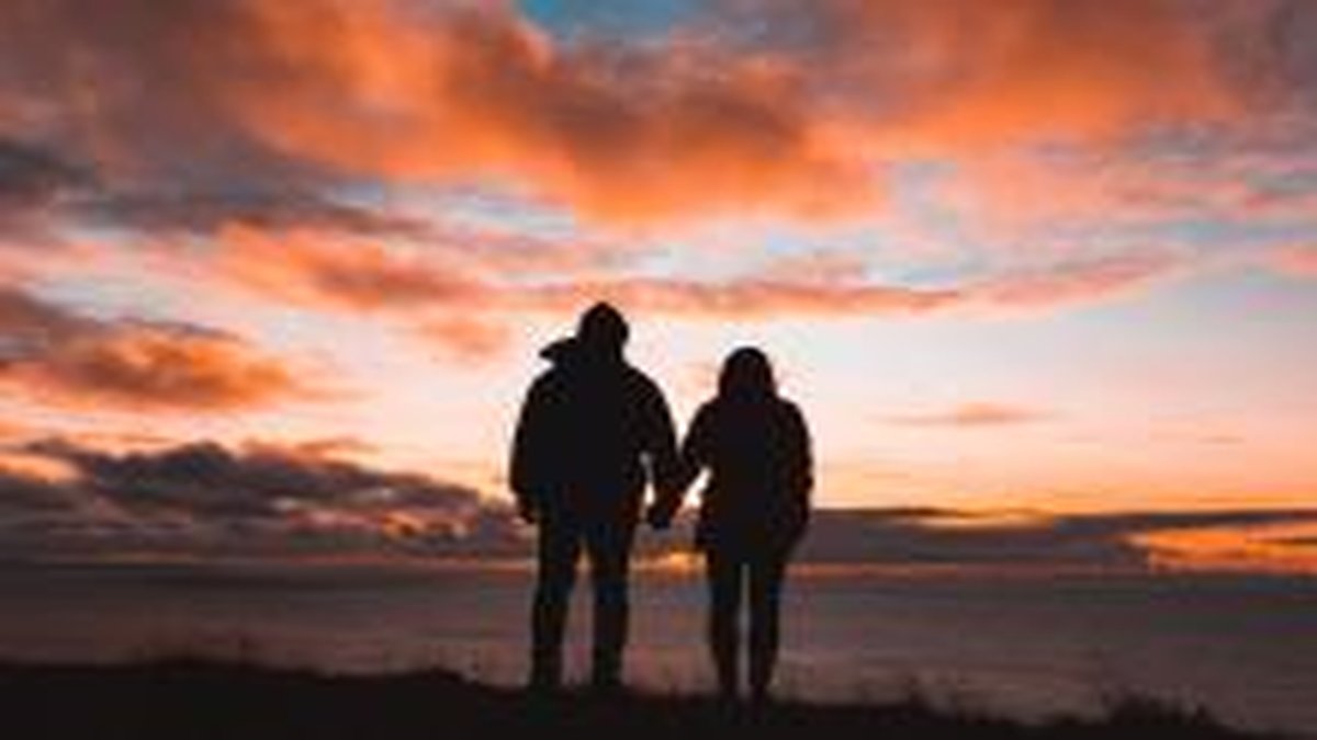 Silhouette of a couple standing side by side, holding hands and looking out over the ocean at sunset, with vibrant orange and pink clouds filling the sky.