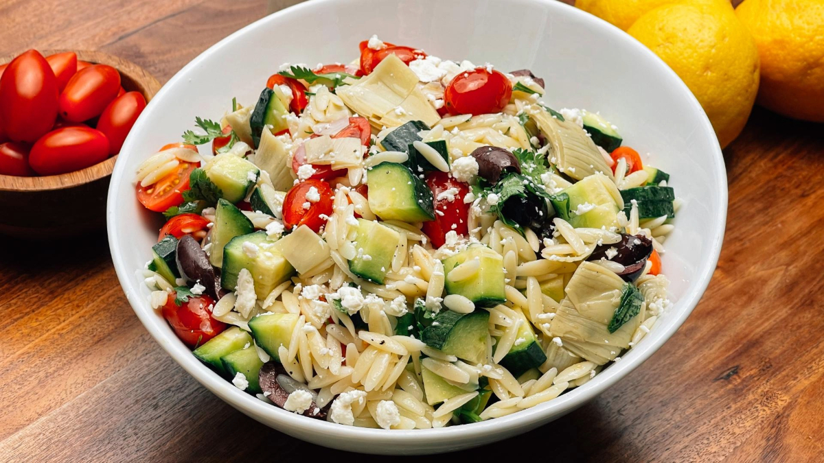 A bowl filled with pasta salad, featuring orzo, cucumbers, cherry tomatoes, black olives, and feta cheese, garnished with herbs. Citrus fruits and a wooden bowl are visible in the background