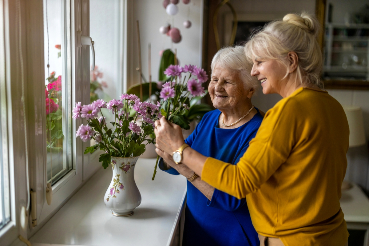 Two individuals are arranging flowers in a vase by a bright window, smiling and enjoying the activity together.