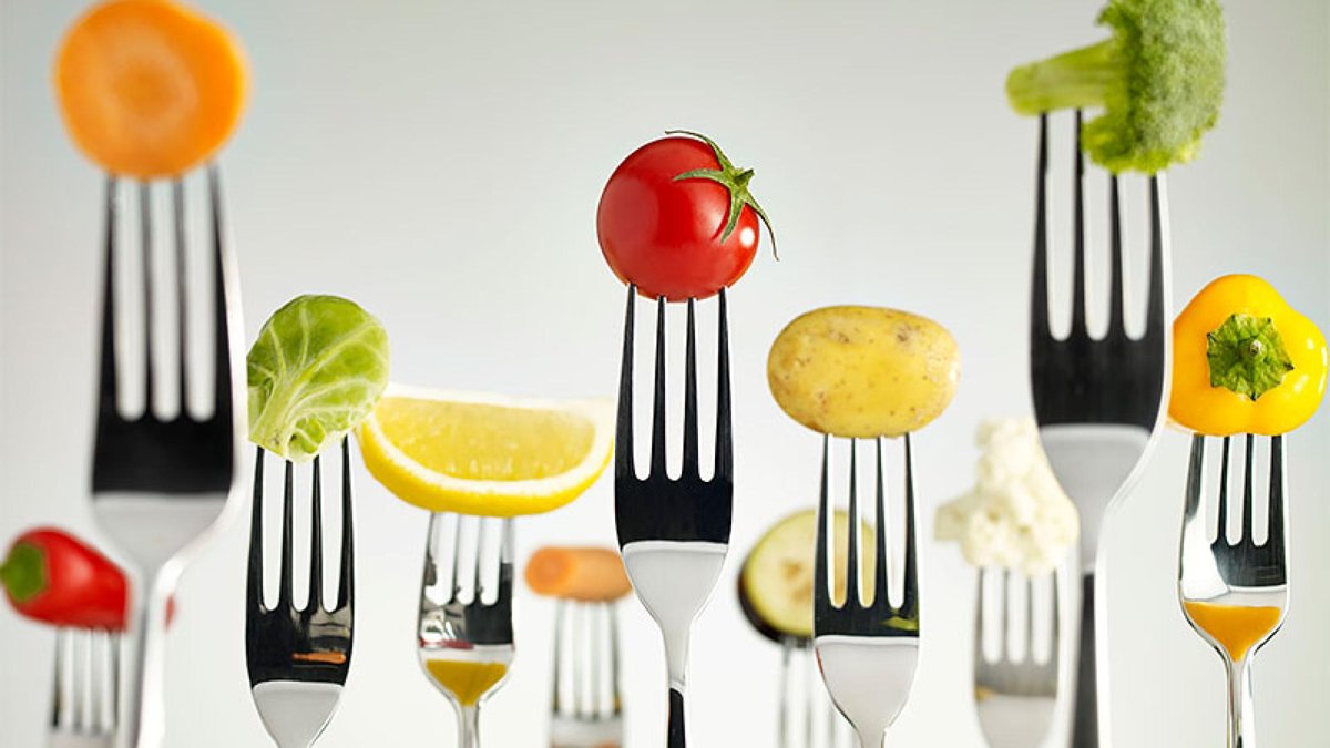 Various colorful fruits and vegetables, including a tomato, lemon, potato, and broccoli, are artfully balanced on forks.