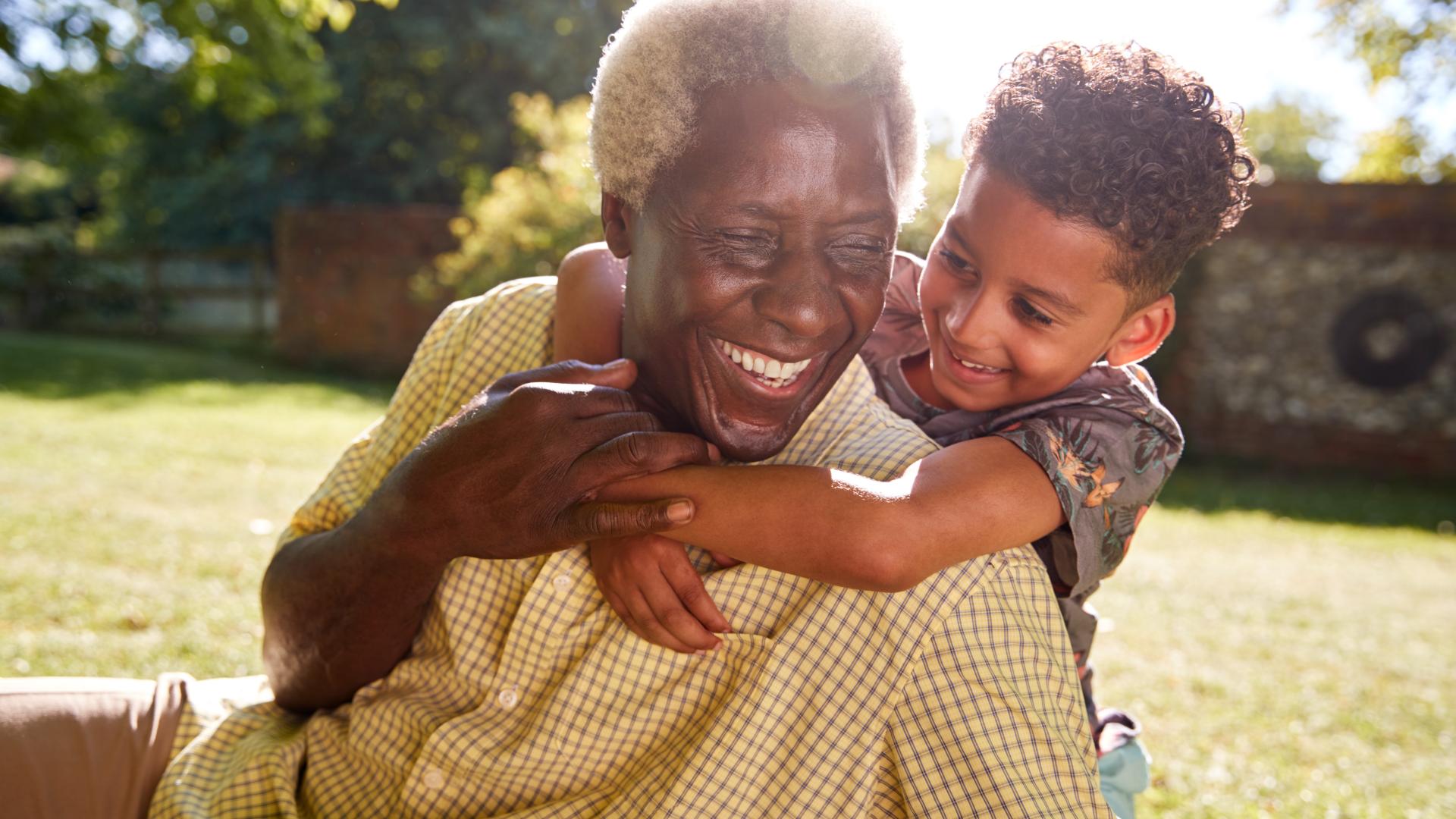 A child hugs an older man around his neck from behind. Both are smiling.