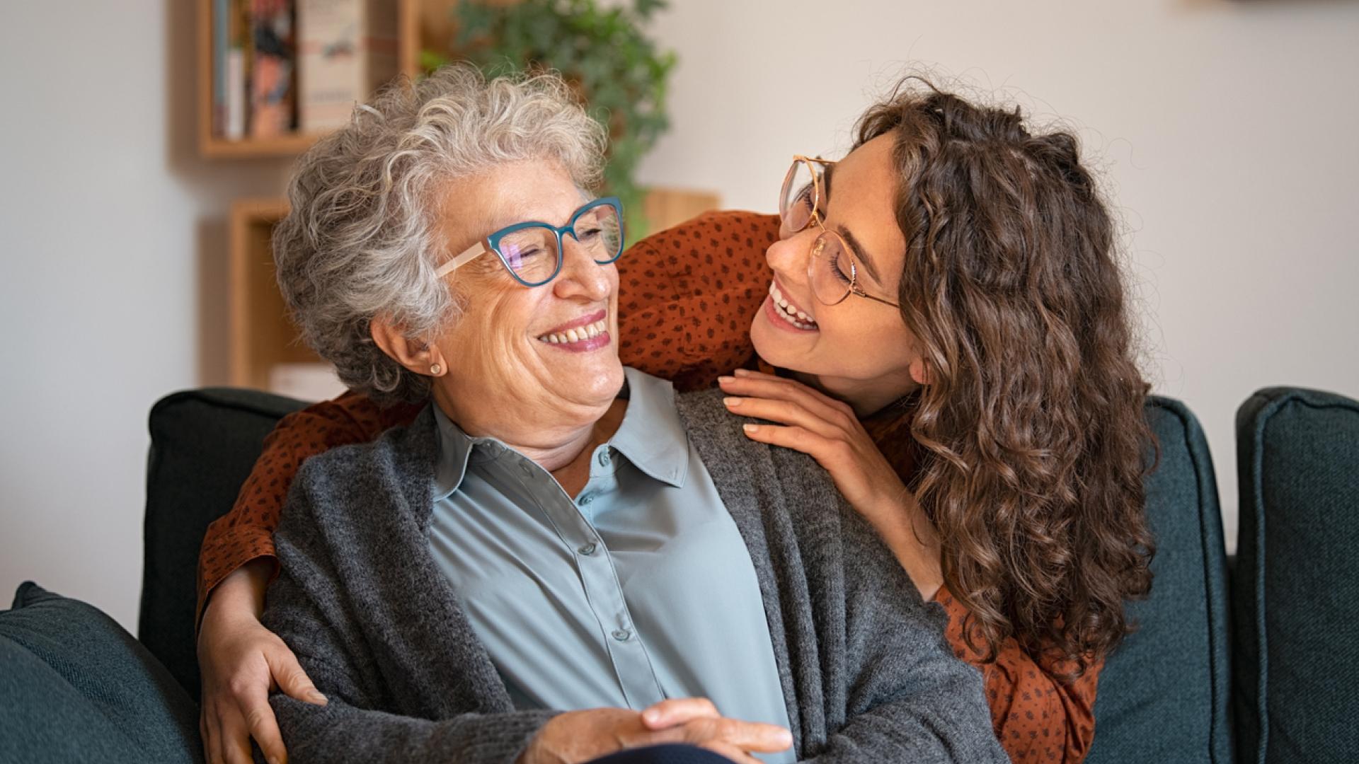 A photograph of a young woman standing behind a seated older woman, leaning in to give her a partial hug. Both women are looking at each other and smiling.