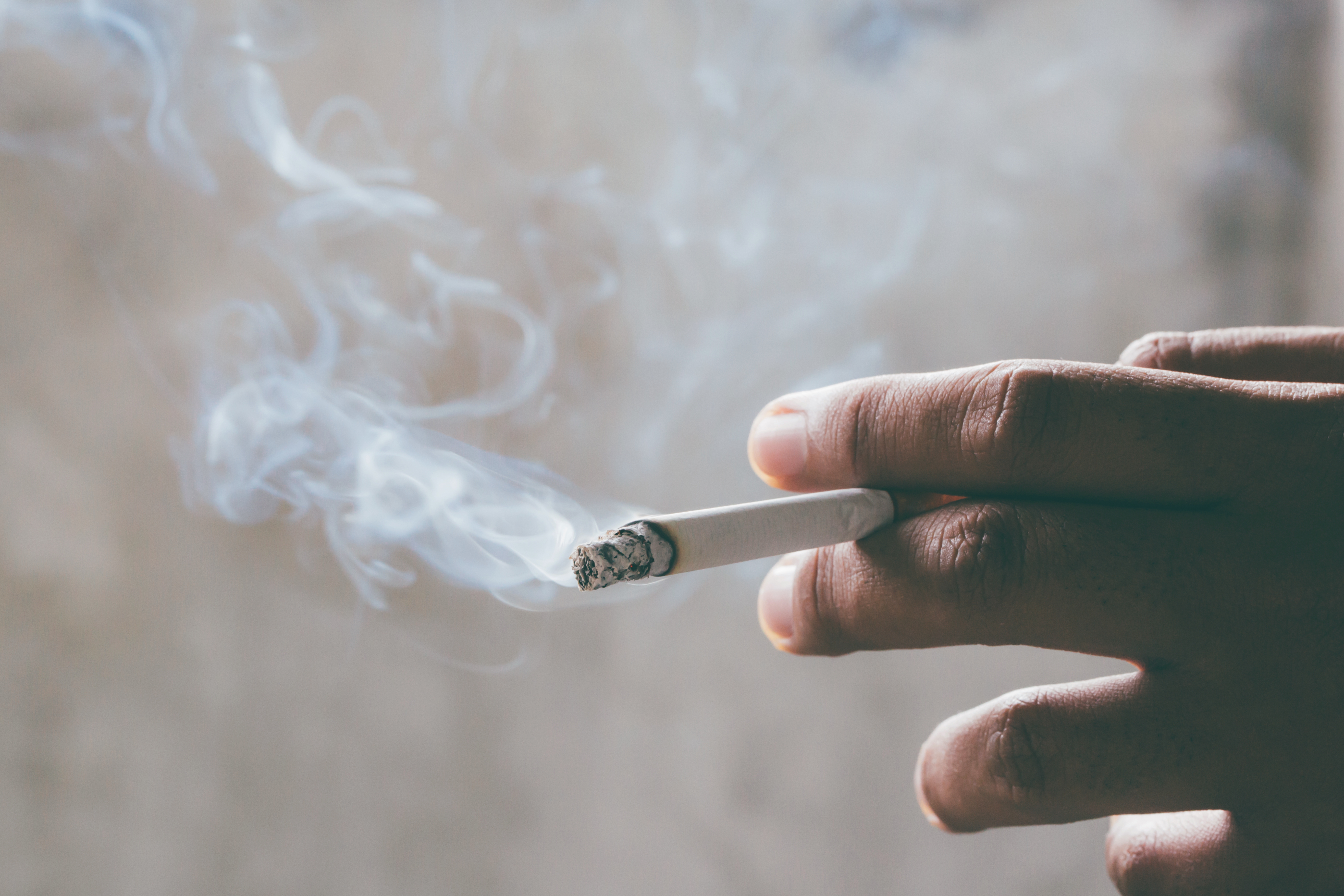 a hand, visible from fingertip to knickle from the back, holds a lit cigarette against a blurry, light-colored background