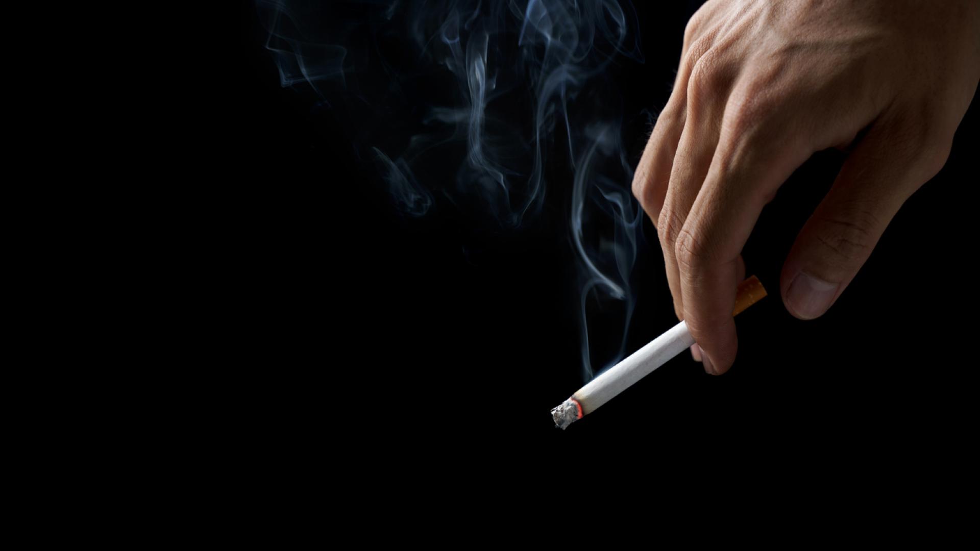 A photo of a hand holding a lit cigarette against a black background.