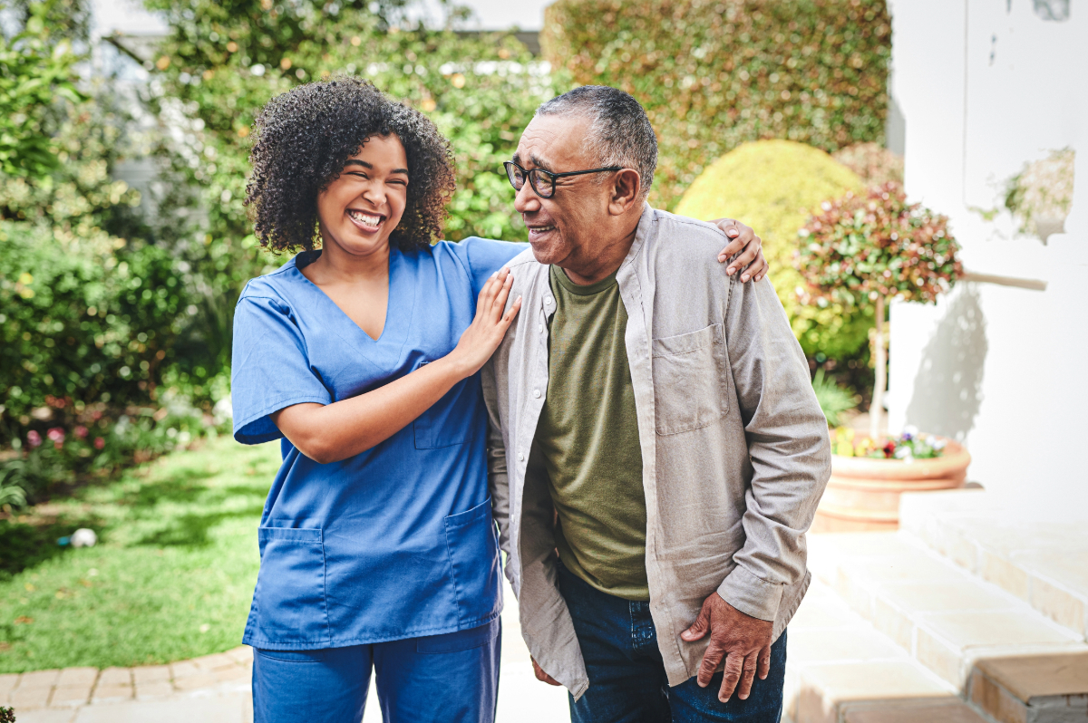 A healthcare professional in blue scrubs smiling and assisting an elderly individual with walking in a garden.