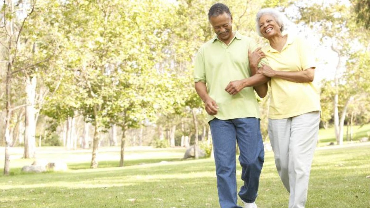 An elderly couple smiling and holding hands as they walk together in a sunny park, surrounded by trees and greenery, enjoying a peaceful and active moment outdoors.