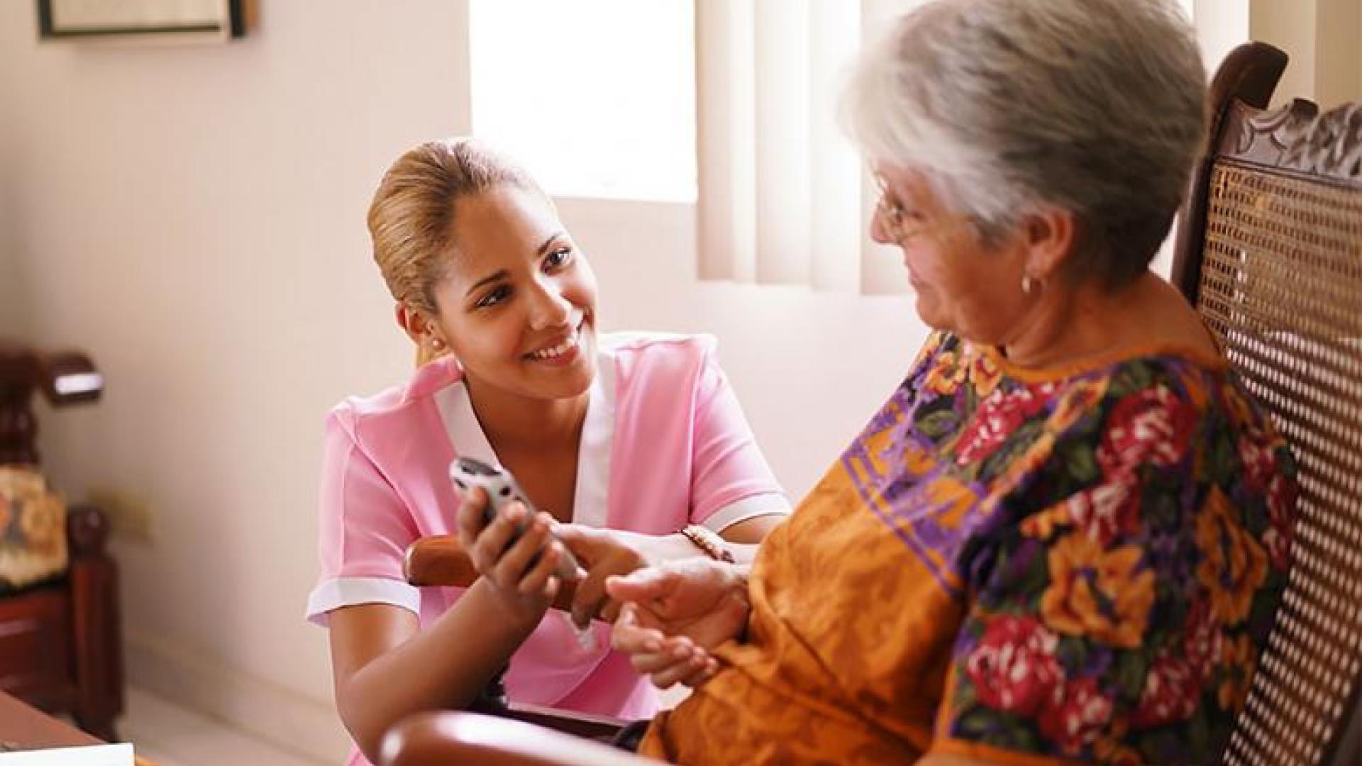 A female aid smiling and helping a senior woman with a digital device.