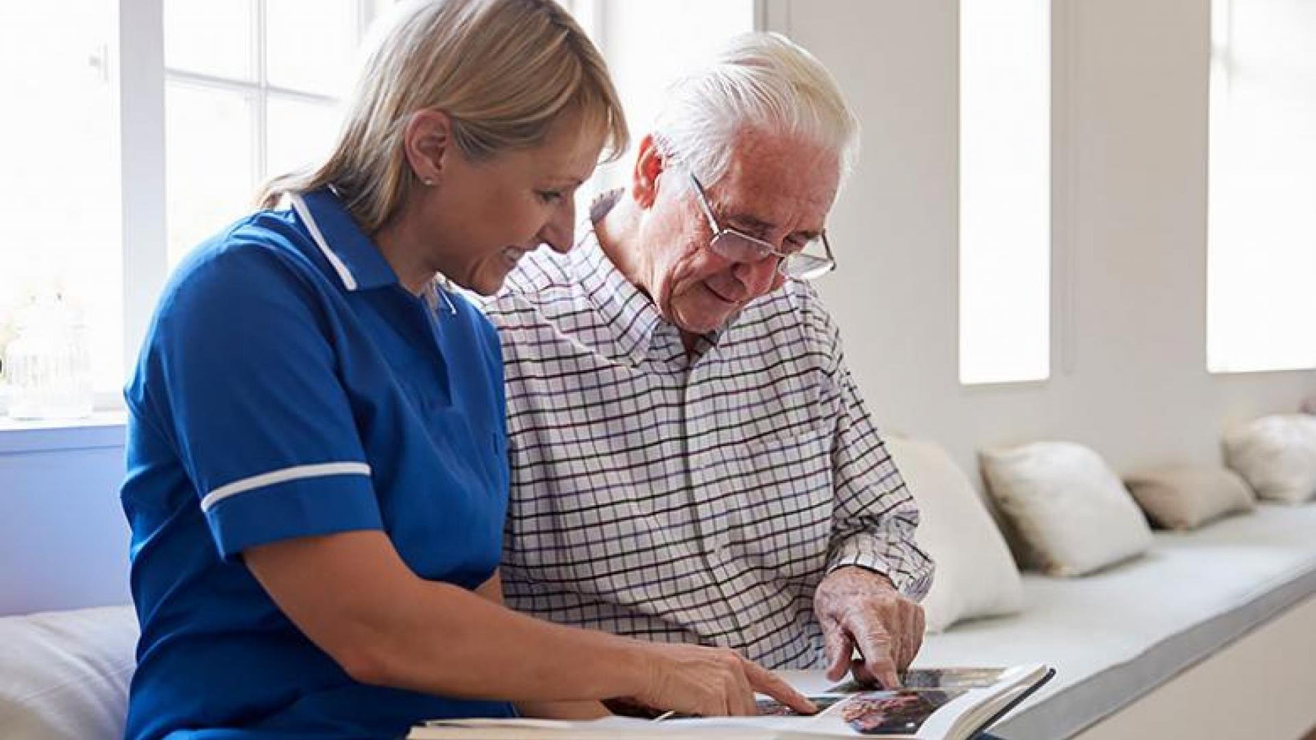 A caregiver in a blue uniform sits with an elderly man, looking through a photo album together in a bright, comfortable room.