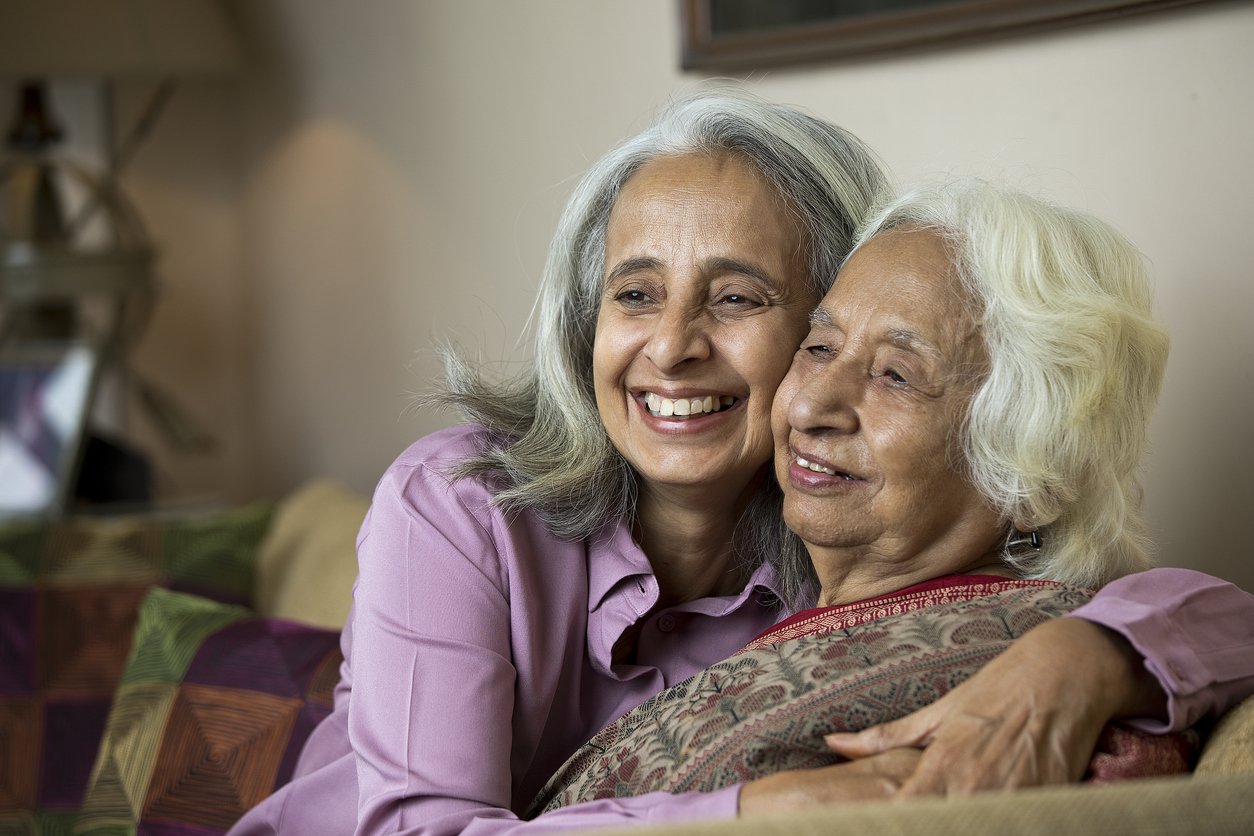 A smiling middle-aged woman embraces her elderly mother as they sit together on a couch.