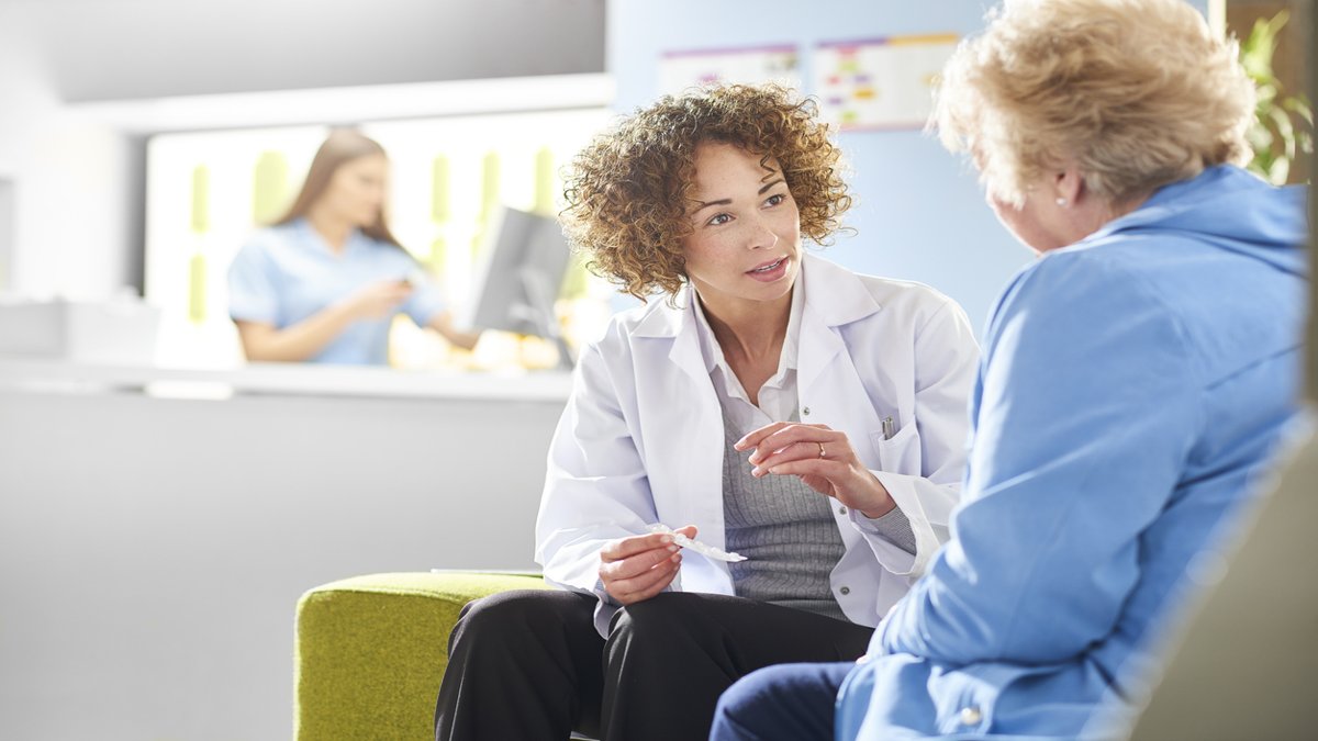 A female doctor with curly hair, wearing a white coat, is sitting and attentively talking to an elderly woman. The doctor holds a prescription in her hand, while the elderly woman listens. A healthcare worker is visible in the background working at a computer.